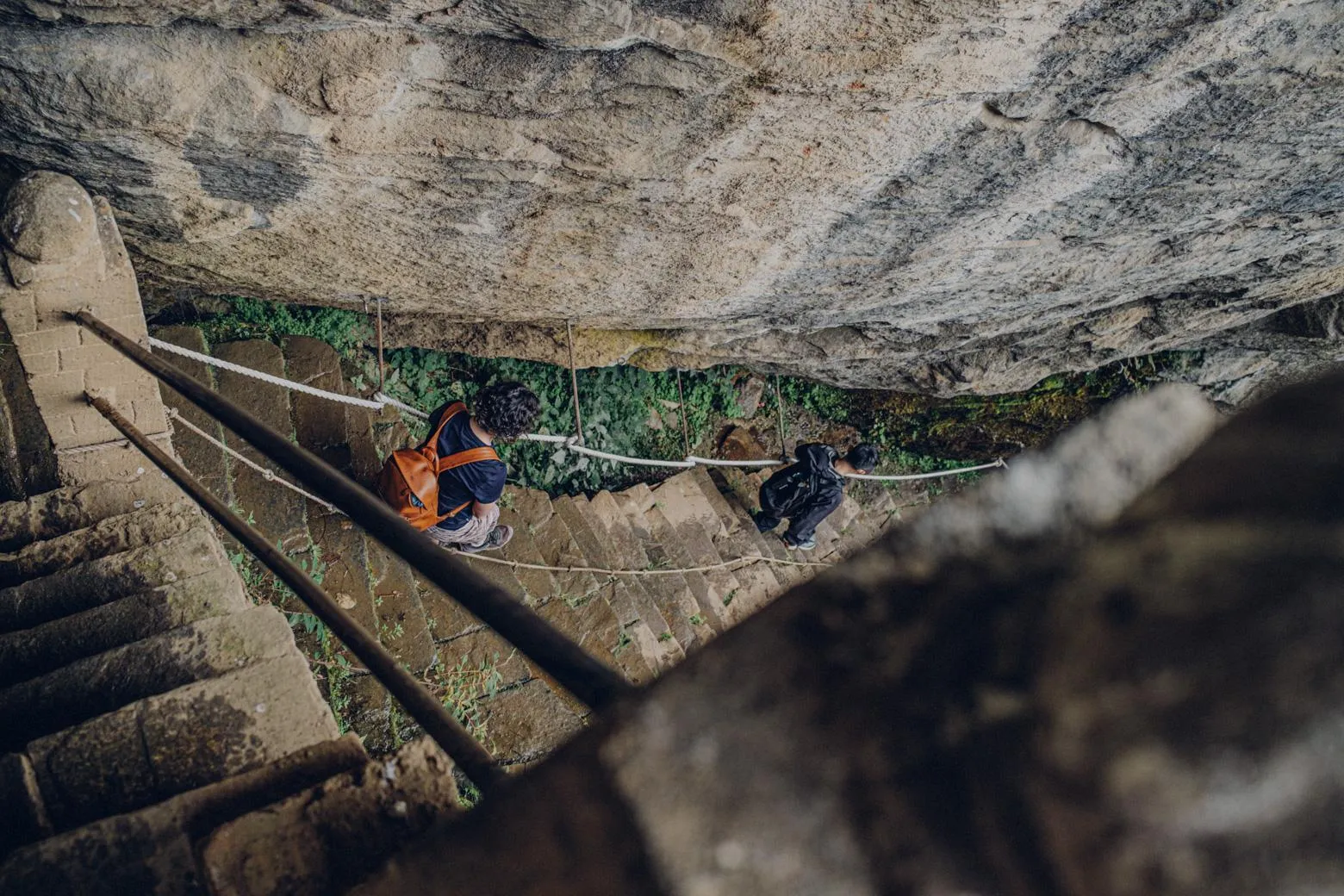 Two travelers descending a rope-lined stone stairway beneath a rock overhang