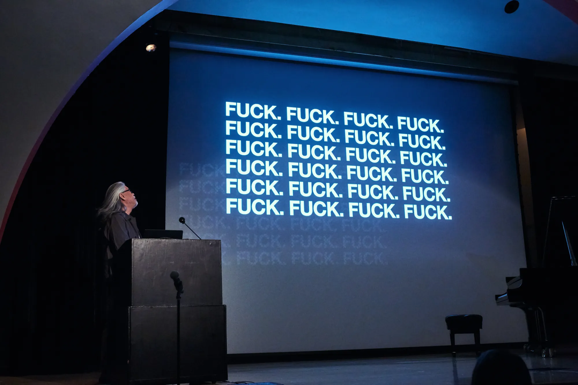 A speaker at the lectern beside a bold projected slide at Behind the Scenes