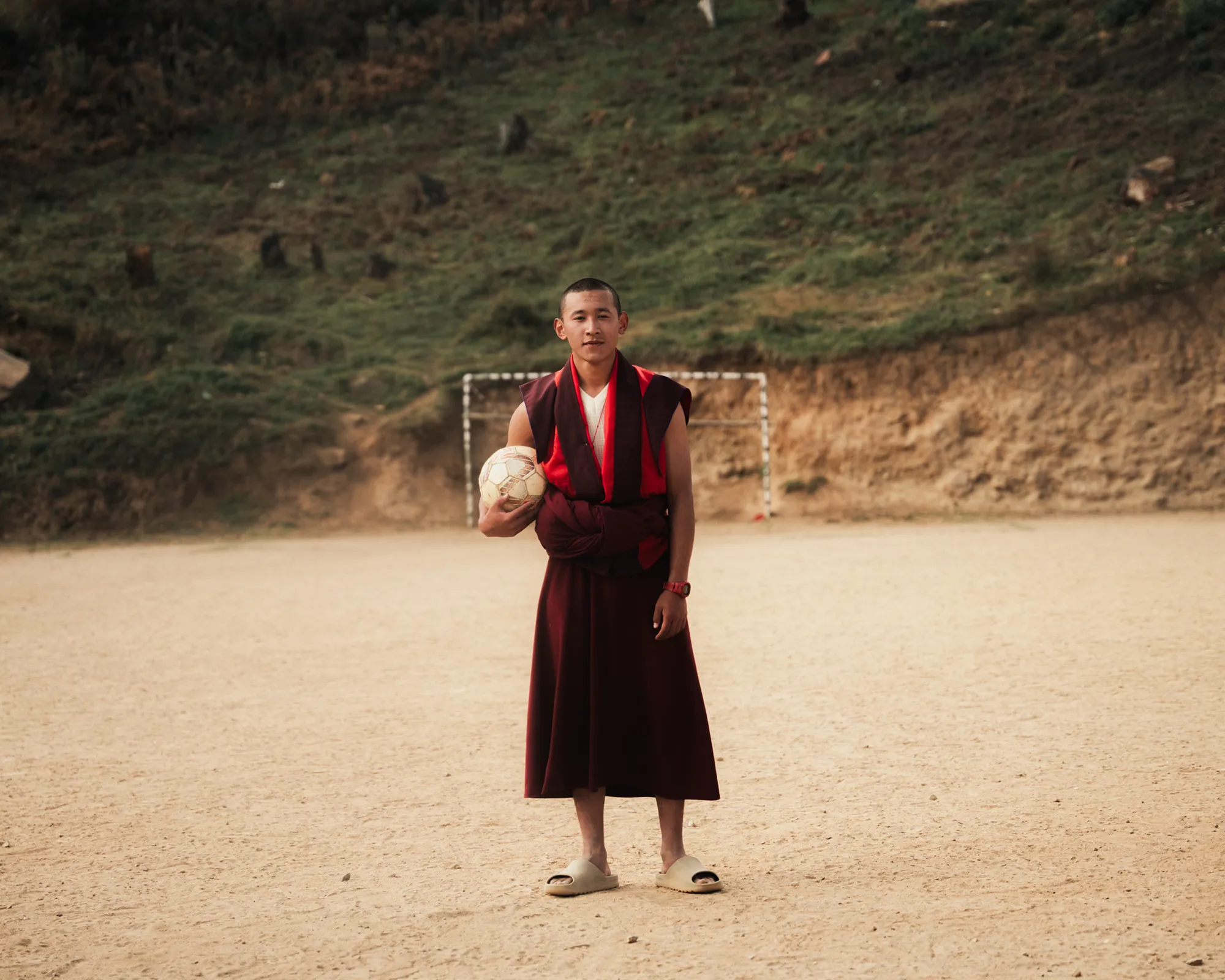 A person in red robes holding a football on a dirt pitch in Bhutan